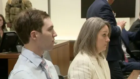 Tyler Robinson is flanked by people while seated in a Provo, Utah, courthouse for his in-person review hearing at the 4th District Court House.