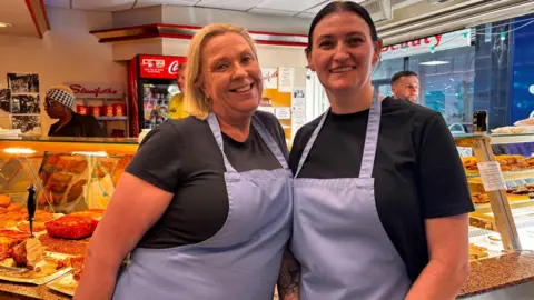 Simon Thake/BBC Two women in purple aprons smile infront of a glass display of cooked food