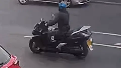 A person wearing a blue helmet and dark clothing rides a black moped along a road, with cars visible nearby.
