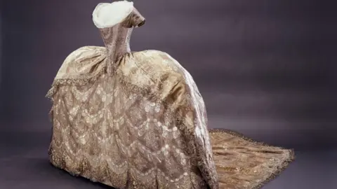 A luxurious court gown in gold and silver tones, featuring an extremely wide skirt supported by panniers, detailed with lace and metallic embroidery, displayed without a head or arms on a mannequin.