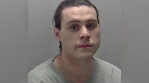 A mugshot of a man who has pale skin and dark hair that is tied back. He is wearing a grey t-shirt.