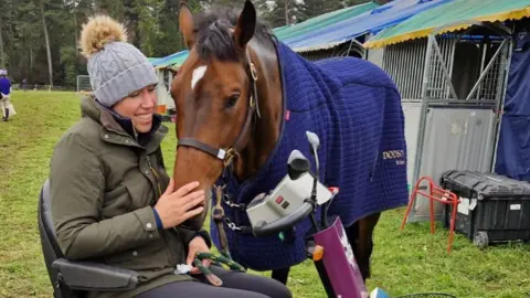 Instagram/Carolinemarchhorses Caroline March in a green waxed jacket and grey bobble hat, who is sitting in a mobility scooter while stroking the nose of chestnut coloured horse. The horse is wearing a blue coat 