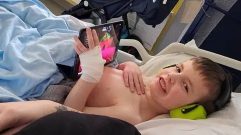 ELHT Arlo lying on a hospital bed under a pale blue blanket smiling, wearing headphones and holding a tablet computer