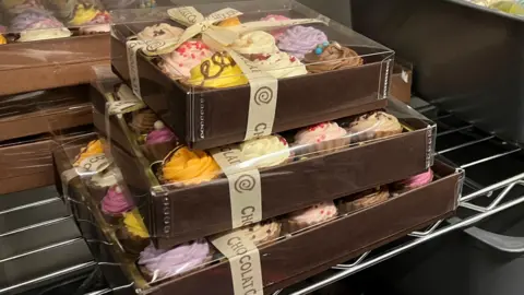 Tom Jackson/BBC Three boxes of chocolates, which are different sizes, are stacked on top of each other on a silver stand. They all are in brown boxes and has beige ribbon tied on it.