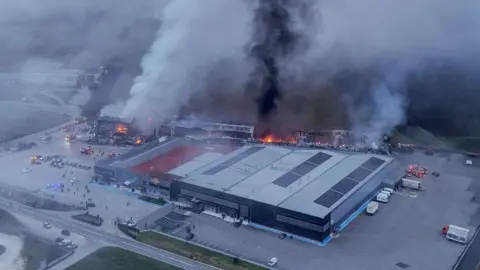 A drone view of the factory with the back of the building in the foreground in tact, but the rear with orange flames and thick black smoke billowing into the sky and filling the screen