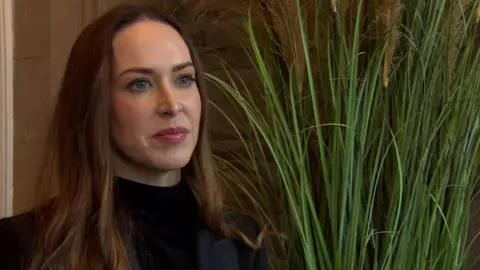 Dr Caitriona Monaghan has long brown hair and is standing in front of a green plant and bricked wall she is wearing a black turtle neck jumper and a black jacket