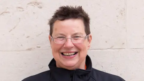 Brass Bands England A woman with short brown hair and glasses smiles at the camera. She is standing in front of a white wall and wearing a black jacket.