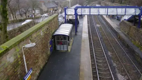 A view from the bridge at Greenfield station. The image shows a second bridge and a small waiting area on the platform beside the railway line, as well as a sign that reads 'Greenfield'. 