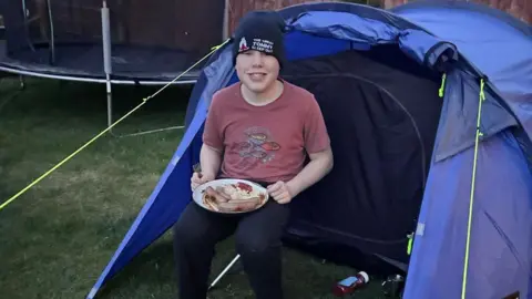 A boy sitting in a camping chair next to blue tent with a plate of food on his lap.