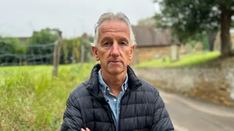 Kate Bradbrook/BBC Nick Frampton with short white hair wearing a black raincoat and standing on grass in front of a church