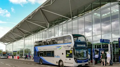 People queueing for bus outside Bristol Airport, which has big glass windows and other buses in the distance.