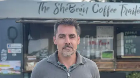 A man with grey hair is standing in front of a coffee cart. He is wearing a green shirt and a grey fleece with a zip at the front.