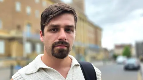 Paul Moseley/BBC Alex Catt is wearing a short-sleeve, cream top. We can see a black bag strap over his left shoulder. Behind him is Norwich City Hall.
