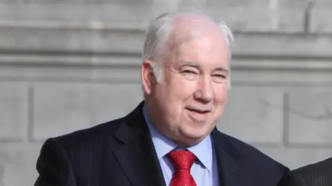 A man with grey hair in a dark suit, light blue shirt and red tie smiling with a wall in the background