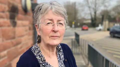 Grey-haired woman wearing rimless glasses and blue cardigan standing in between a brick wall and black railings. A car park can be seen in the background.