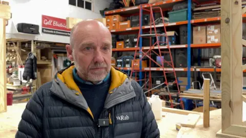 Martyn Maguire wearing a jacket, stood in a workshop. He is bald and has grey stubble.