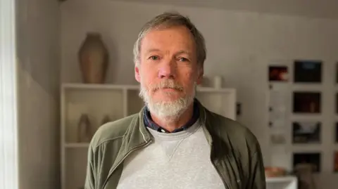 A man with a beard, in a grey jumper and green jacket, stands in front of some pottery on shelves.