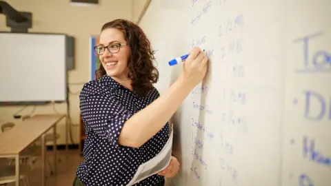 Getty Images A happy curly brown haired female teacher writing on whiteboard in classroom. She has a navy polka dot shirt on 