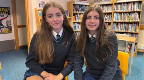 Two pupils at John Cabot Academy, sitting on yellow chairs in the library. They are both wearing a school uniform consisting of a blue and black checked skirt, navy jumper and dark tie. Both have brown hair and are sitting close together.