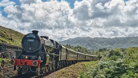Eugene Rutter A vintage black steam locomotive pulls passenger carriages along a railway line through a lush green landscape, with mountains and dramatic clouds in the background.