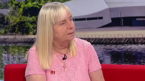 Margaret Aspinall has long blonde hair and is wearing a pink t-shirt with a red 97 pin badge and a gold crucifix. She is sat on a red sofa as she is interviewed on BBC TV.