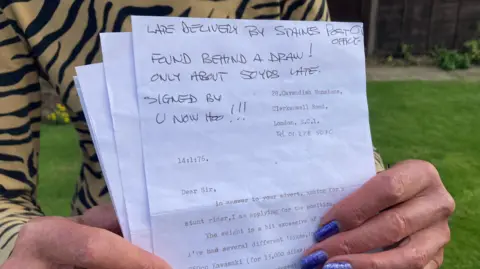 Tizi Hodson holds up the letter found by a post office after 50 years. A note written in black pen explains that it is a "late delivery by Staines Post Office" and that it was found behind a drawer, only about 50 years late.