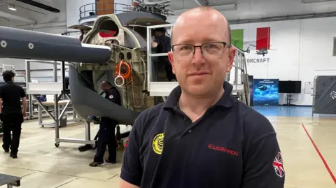 Tom Spencer, one of the team on the project, standing in a hangar with the unfinished body of a Proteus helicopter in the background.