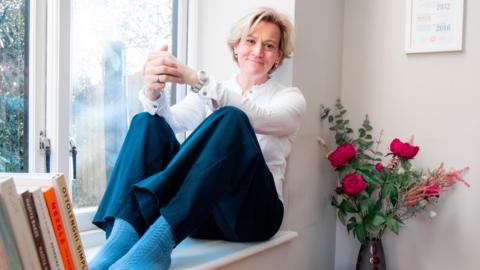 Picture shows Anna Doneghey wearing a white shirt and dark trousers, sat in a large window seat besides a vase of flowers and a selection of recipe books.  