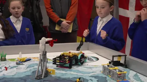 BBC Lots of school pupils crowded around tables covered in pieces of Lego. 