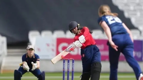MCC/Jed Leicester Afra strikes a cricket ball with her wooden bat. A wicket keeper wearing navy is poised in the background ready to catch the ball.