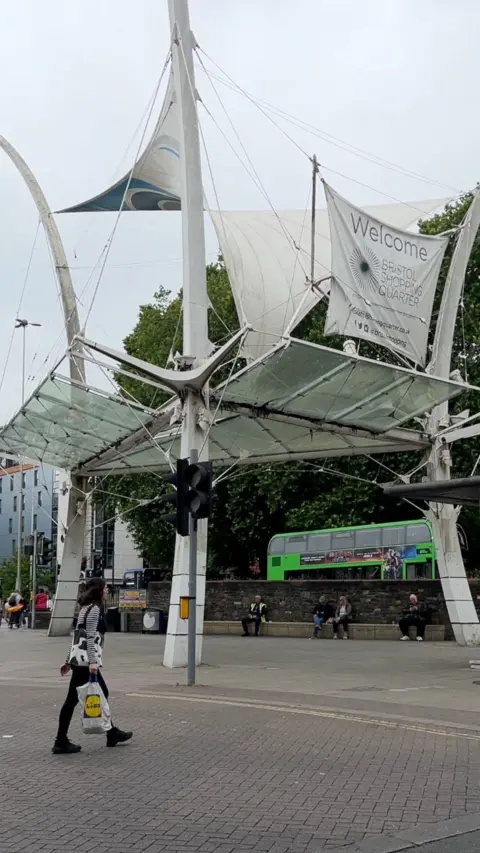A white sail structure placed near a road with shops in the background.