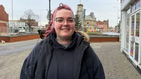 Becki Bowden/BBC Abbie Bircumshaw standing in goole town centre. She is smiling at the camera. She has pink hair, tattoos on her neck and piercings on her nose and lip. A clock tower can be seen behind her.