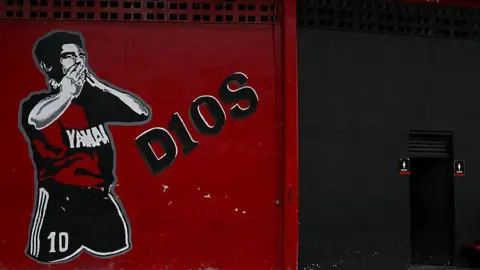 Getty Images A red and black mural on a wall depicting Diego Maradona kissing his hands. Text on the mural reads D10S.