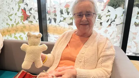 Resident Jennie Rogers, looking at the camera and smiling, in a cream cardigan and orange t shirt, holding a cream bear she has knitted.