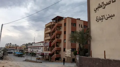 Brown buildings are seen under a cloudy sky, with a small figure walking past and debris on the ground