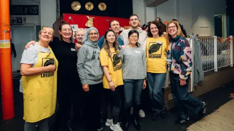 Zohaib Alam/Lifeshare Diogo Dalot stands among a group of women plus one man. They have their arms around each other and are smiling. Some wear yellow t-shirts or aprons that read 'Lifeshare'