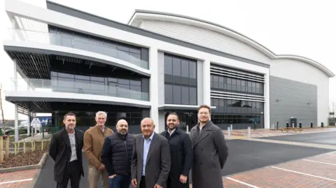 Chris Kirkland, City of Wolverhampton Council, Alex Morgan, from Stoford, Kunal Patel, Ravi Patel and Nikul Patel all from Task and councillor Chris Burden, City of Wolverhampton Council stand outside the new premises. They are in the car park in front a large white and grey glass building.
