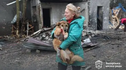 Zaporizhzhia city council A woman carries a dog with the backdrop of a damaged building