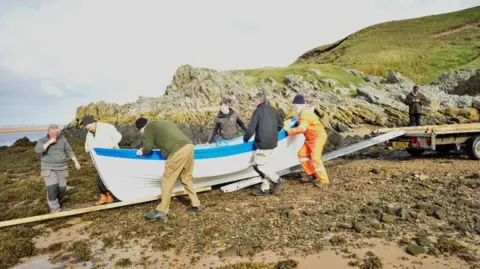 Strathnaver Museum People push the white and blue boat down a ladder and wooden plank on a rocky beach.