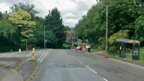A Google Street View image of the junction of Redbrook Road and Beever Lane. To the right of the road is a bus stop. At the bottom of the hill a few houses are visible through the trees.