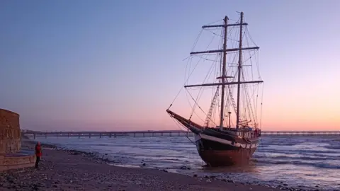 Clint Kinrade A traditional tall ship sits upright in the surf of beach with the tide in, run aground. It is sunset with a orange horizon blending into a darker blue sky. There is a long iron pier in the distance. 