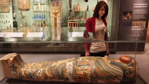 Getty Images A woman stands in an exhibit full of Egyptian artifacts at Oxford's Ashmolean Museum. She is looking down upon a glass display case that contains a decorated sarcophagus.