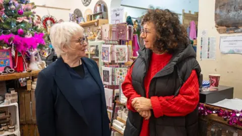 Northamptonshire OPFCC Danielle stone with short white hair and glasses, wearing a dark top and jacket, talking to Julie Teckman with long curly dark hair and glasses, wearing a red sweater with black gilet. They are standing in a shop with cards, flowers and other items for sale.