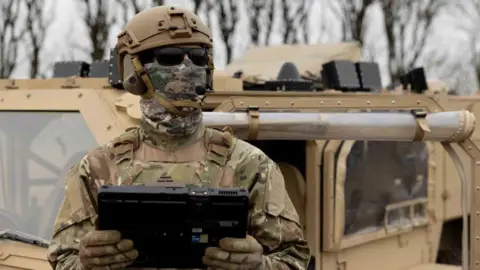 MyDefence A soldier wearing a helmet, mask and sunglasses holds a large table computer and looks upwards. Behind him is a military vehicle.