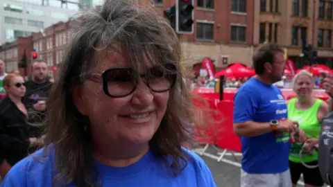 A woman wearing a blue T-shirt and sunglasses and with long brown and grey hair is smiling while standing next to other runners.