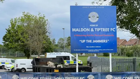 The outside of Maldon & Tiptree FC. There is a sign at the front of the grounds for the Drewitt-Barlow Stadium. The sign is blue with white writing and says, HOME OF MALDON & TIPTREE FC. A Welcoming Hub For The Local Community. "UP THE JAMMERS". In the background there are a number of police vehicles parked up.