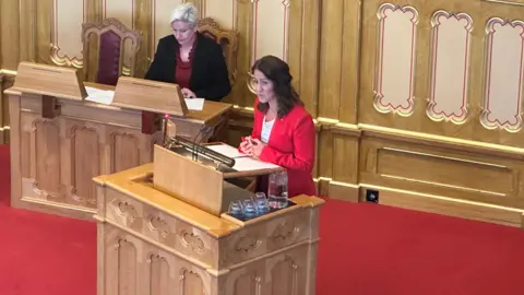 Tonje Brenna speaks at a large wooden lectern. She has long brown hair and is wearing a red suit