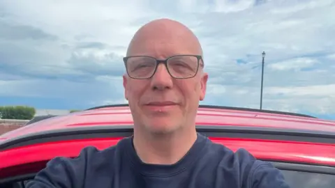 Supplied A selfie of a man wearing a navy jumper and glasses. He is standing outside, seemingly in front of a red car. 