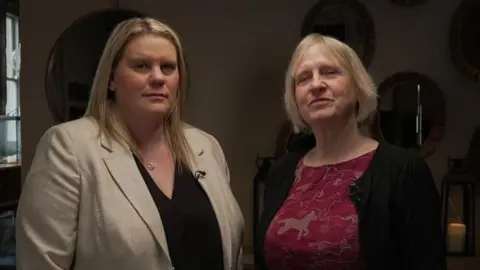 Micala and Sue stand side by side looking defiant. Micala has long blond hair and is wearing a black shirt and light brown jacket. Sue has short blond hair and is wearing a red top with brown horses on it and a black jacket. Behind them is a wall with several mirrors on it 