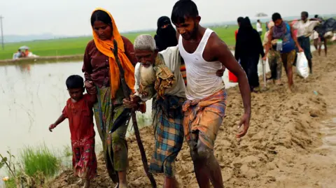 Rohingya refugees walk on the muddy path after crossing the Bangladesh-Myanmar border in Teknaf, Bangladesh, September 3, 2017. A man and a woman in an orange scarf support an elderly man with a long white beard who is using a stick to walk. The woman clutches a little boy's hand in her free hand. Green fields, a pond and hills can be seen in the background
